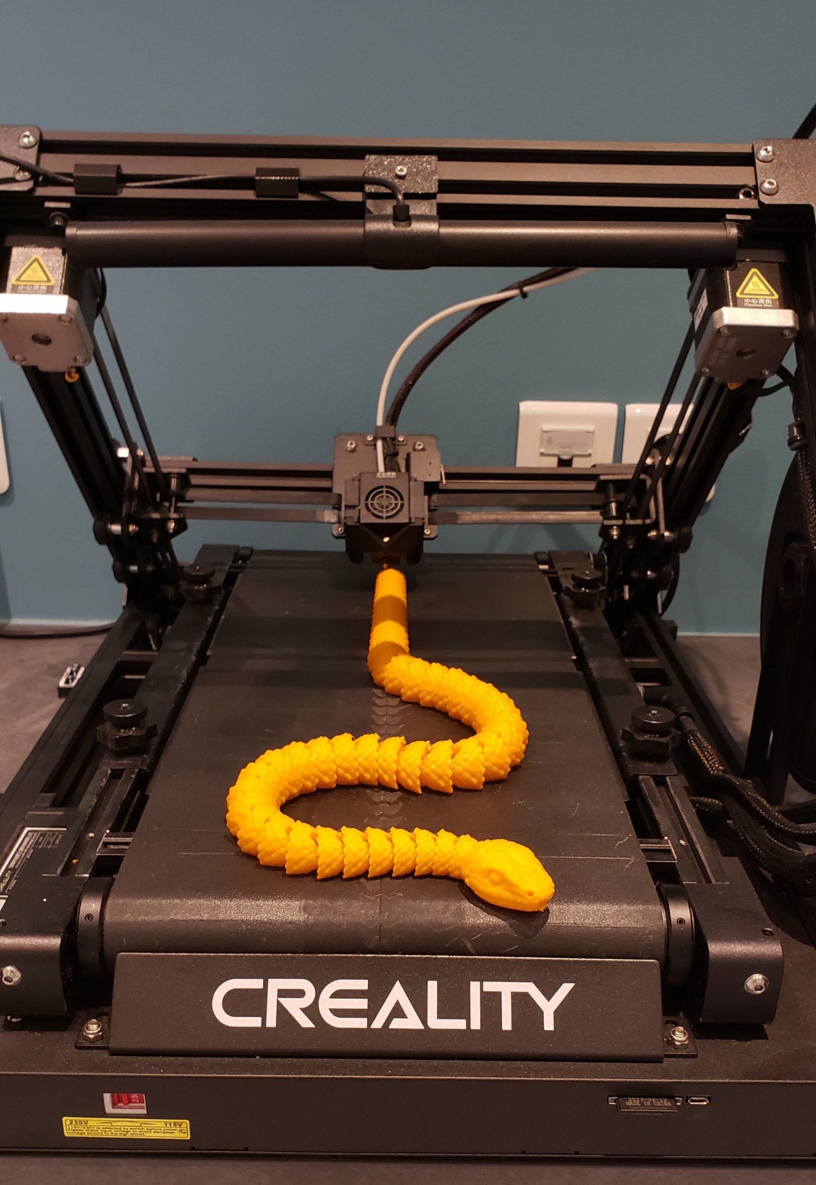 Gros plan sur une imprimante 3D avec un Z infini qui est un tapis roulant. Sur l'imprimante, un serpent articulé très long réalisé grâce à cette machine.